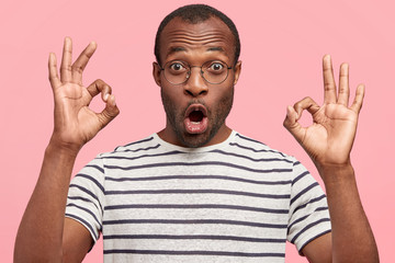 Emotional surprised dark skinned male shows ok sign with both hands, demonstrates that everything is fine, shows his approval, gestures against pink wall. Body language and facial expressions