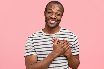 Photo of satisfied dark skinned male keeps hand on chest, wears casual t shirt, expresses his politeness, devotion and friendly manners, being touched by something, poses against pink packground