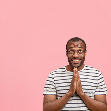 Vertical shot of pleasant looking delighted male holds hands in praying gesture, looks hopefully upwards, asks for favour or mercy, wears glasses and casual striped t shirt, stands against pink wall