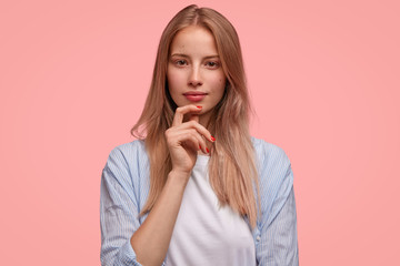 People, beauty and youth concept. Good looking young woman has serious expression, listens interesting conversation with great interest, holds chin, dressed in casual outfit, poses against pink wall