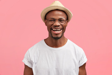 Middle aged dark skinned hipster in stylish hat, giggles and smiles gladfully at camera, being in...