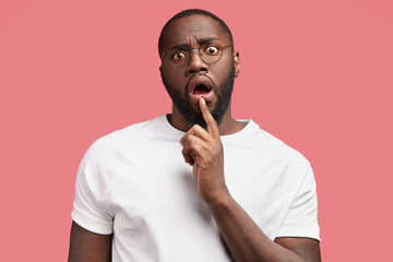 Indoor studio shot of attractive stupefied African American male with stubble, opened eyes, stares at camera with shocked expression, recieves unexpected task, poses against pink background.
