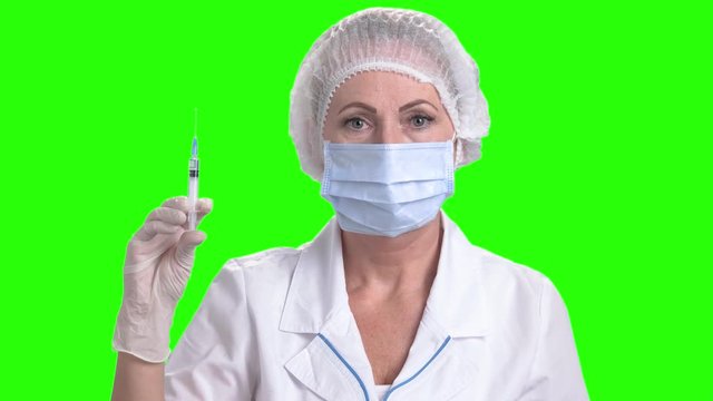 Doctor woman holding syringe on green screen. Portrait of confident medical worker in cap and mask holding an injection on Alpha Channel background.
