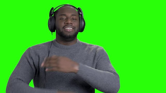 Man in headphones is dancing on green screen. Cheerful energetic african-american guy is listening to music, dancing and showing thumbs up on chroma key background.