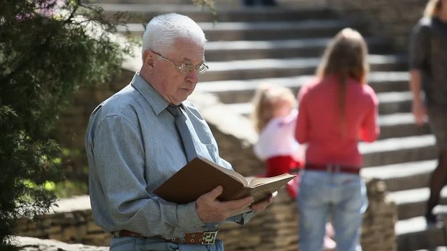 Portrait of a spic and  span man in a grey shirt and glasses sitting on a parapet and reading a book near a stony staircase with people in summer