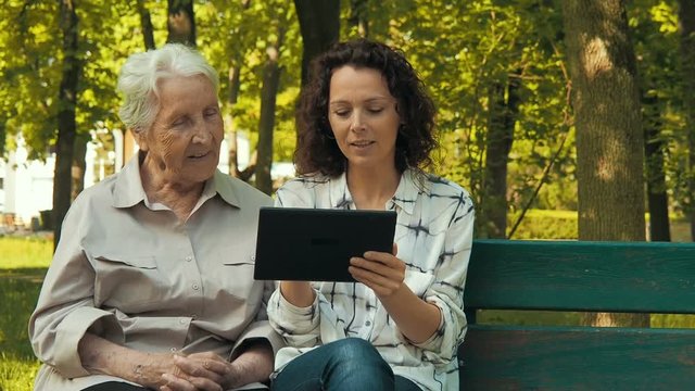 Look at the pictures on the tablet. An elderly woman with a tablet. A woman with an elderly mother with a gadget in the park.