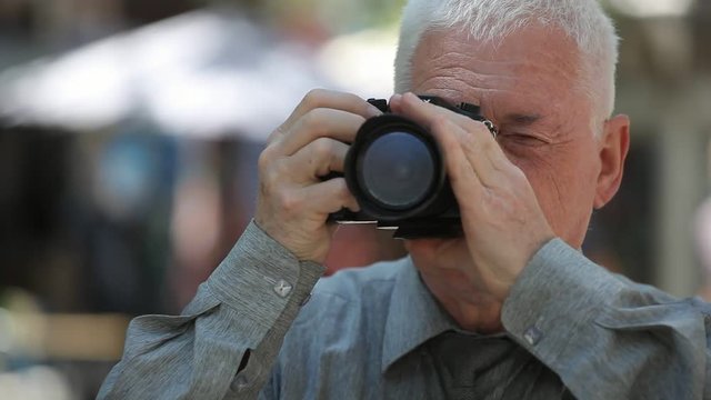 Bokeh shot of a grey-headed man with a crew haircut in a grey shirt standing in a street, raising his camera and taking photos in summer 