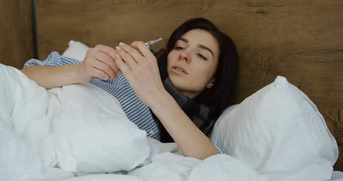 Close up of the Caucasian unhealthy woman lying in the bed with a termometer as measuring a temperature and feeling herself bad. Inside