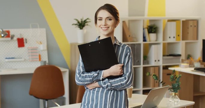 Portrait shot of the young pretty woman with a black folder in hands standing and posing in the modern office room. Indoors