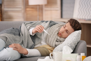 Sick young man with thermometer suffering from cold on sofa at home