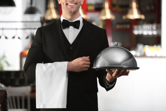 Waiter in elegant uniform holding metal tray and cloche at workplace