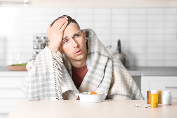 Sick young man eating broth to cure cold at table in kitchen