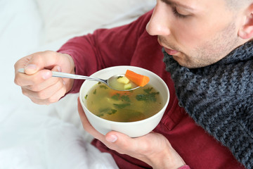 Sick young man eating broth to cure cold in bed at home