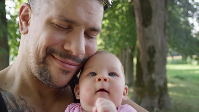SLOW MOTION, CLOSE UP, COPY SPACE: Big tattooed father sharing priceless gentle moments with his baby girl in the green park. Cheerful muscular dad kisses his adorable newborn daughter on the cheek.