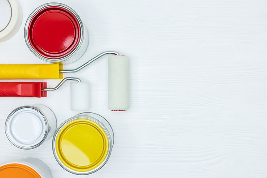 cans with white, yellow, red and orange paints, paint rollers on white wooden background. flat view