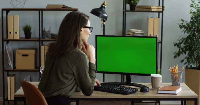 Rear view on the caucasian woman sitting at the office desk and taping on the keyboard of the computer with green screen, thinking while texting. Chroma key. Indoors
