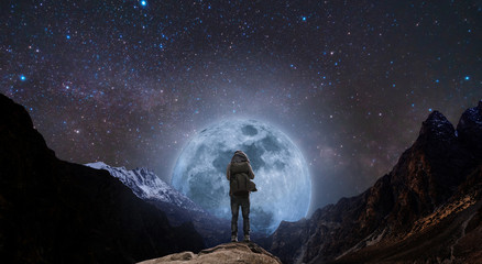 a man with backpack standing on mountain peak at night, and silhouette mountain with bright full...