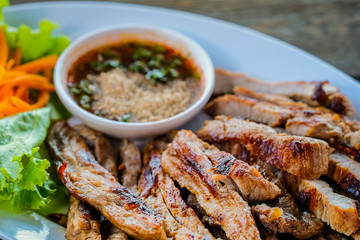 Grilled pork in a white dish with vegetables and dipping sauce on a wooden table.