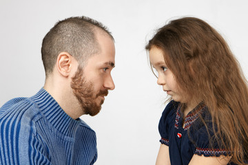 People, family, parenthood and bringing up concept. Sideways shot of handsome young man with stubble posing at white wall with his little daughter, looking at each other, having some argument