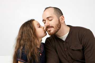 Family bonds, love and relationships concept. Studio shot of cute adorable little girl with long voluminous hair kissing her unshaven father on cheek, showing her gratitude for birthday gift