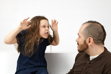 Studio shot of funny emotional little girl with messy hairstyle grimacing and making fearful gesture with both hands, trying to scare her attractive bearded father while playing together indoors