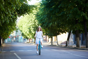 Fototapeta na wymiar Smiling young female in white dress and straw hat riding blue bike down wide beautiful street with no people around just surrounded trees. Vintage turquoise bicycle green street city happy enjoying