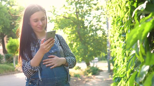 Young pregnant girl dressed in overalls looking at phone, on green background.