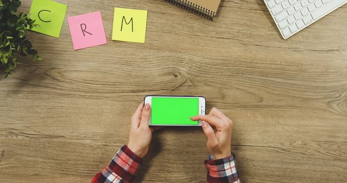 Close up on the woman's hands taping on the white horizontal smartphone with a green screen on the wooden table. SRM. Chroma key.