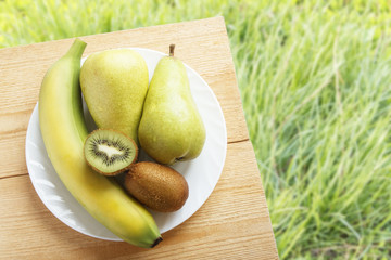 Ripe banana, kiwi fruit and pear on wooden table on background of green grasses. Rustic lifestyle concept. Top view. Copy space