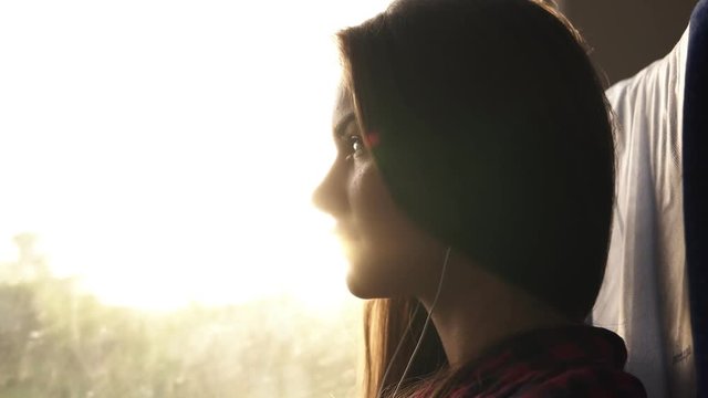 Attractive girl sitting in the shadow in train. Listening to the music with earphones and mobile phone. Sun rays in window. Nature landscape outside