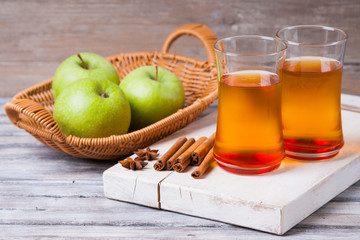 Apple juice in glasses, spices on wooden cutting table and apples in wicker basket 