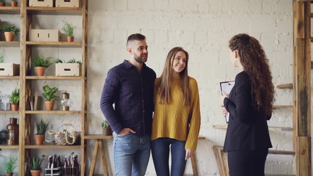 Female realtor in suit is showing loft style flat to young man and woman looking for new apartment. Buyers are excited about interior, they are smiling and talking.