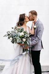 Portrait of a beautiful couple in love on your wedding day. Walking near the house with white walls and greenery. Amazing kisses and embraces of the bride and groom with a bouquet
