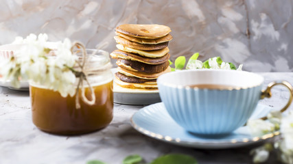A pile of fritters next to a cup of tea and a jar of honey on a gray background among the flowers of the acacia. Spring delicious breakfast