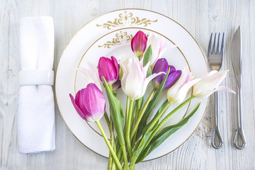 Festive table setting with original cutlery on white napkin and bouquet of tulips on white wooden background top view