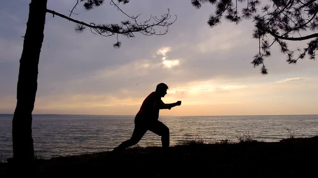 A man practices karate punches on the shore of the reservoir during the sunrise. Silhouette of an adult man practicing strokes with his hands.