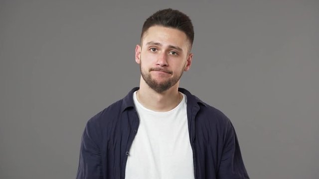 Portrait of indifferent man in casual clothing shrugging and expressing misunderstanding or have no clue with throwing up hands, isolated over gray background slow motion. Concept of emotions