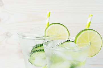 Fresh summer organic drink with slices cucumber, ice, lime, straw on white wood board, closeup.