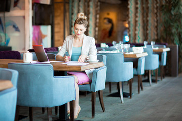 Busy young employee in formalwear sitting in cafe and making notes while waiting for colleague or...