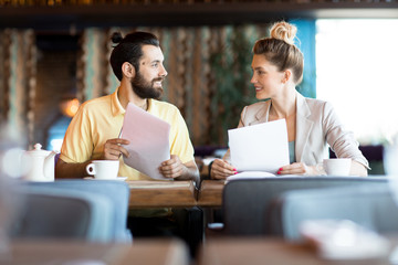 Two young accountants with financial documents looking at one another while sitting by table in modern cafe