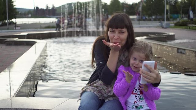 Mother and cute little daughter having fun and doing selfie in the park next to the fountain.