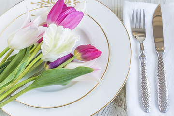 Festive table setting with bouquet of multicolored tulips on white plates and cutlery on white linen napkin