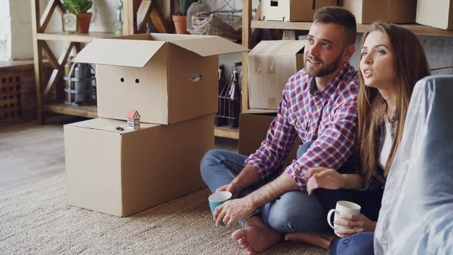Cheerful young woman is talking to her husband about their new house sitting on floor during relocation. Happy people, housing, relationship and real estate concept.