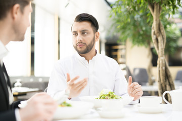 Young manager and his colleague sitting by served table in cafe, having business lunch and discussing some working problems