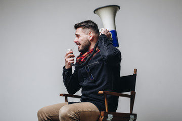 Side view of young attractive movie director shouting on the megaphone while sitting on the chair...