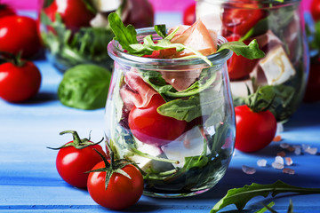 Salad with arugula, cheese, cherry tomatoes and prosciutto in glass jars, pink blue background, selective focus