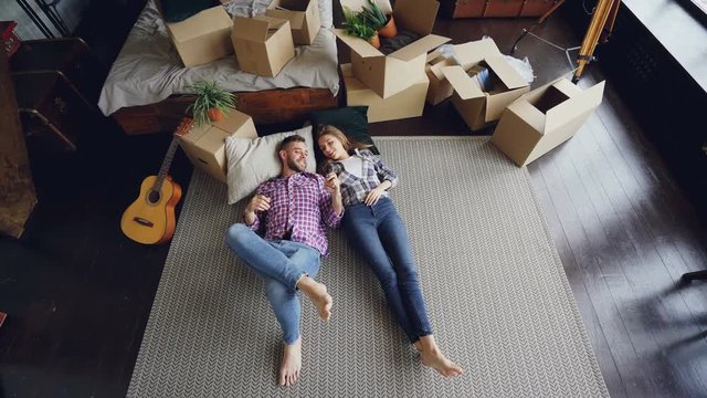 High shot of young couple lying on floor of their new house in bedroom, talking, laughing and holding hands. Carton boxes, guitar, carpet and bed are visible.