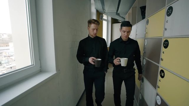 Two male office workers going through corridor with lockers. Concept of: business people, office corridor, management consulting, coffee drinking, locker room, business communication.