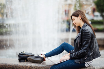 Beautiful teen woman is using her laptop computer outside, while sitting by a fountain in a town square