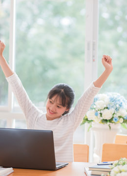Portrait of happy Asian woman manager sitting at her laptop computer in work office and raised hands up with smiling because winner or success celebrating perfect done work.